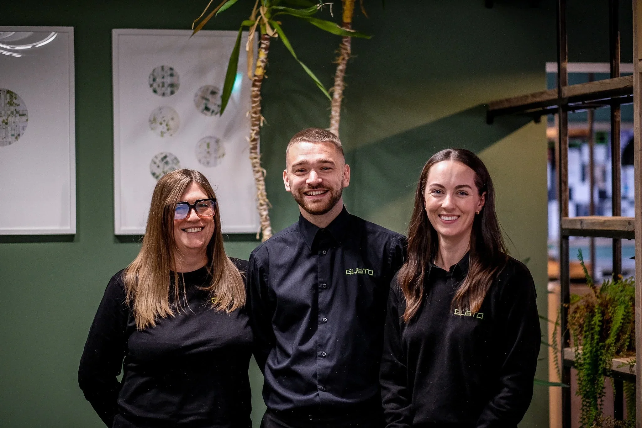 Three people dressed in black shirts with a logo, smiling in a green-themed indoor setting with art circles on the wall and plants around.
