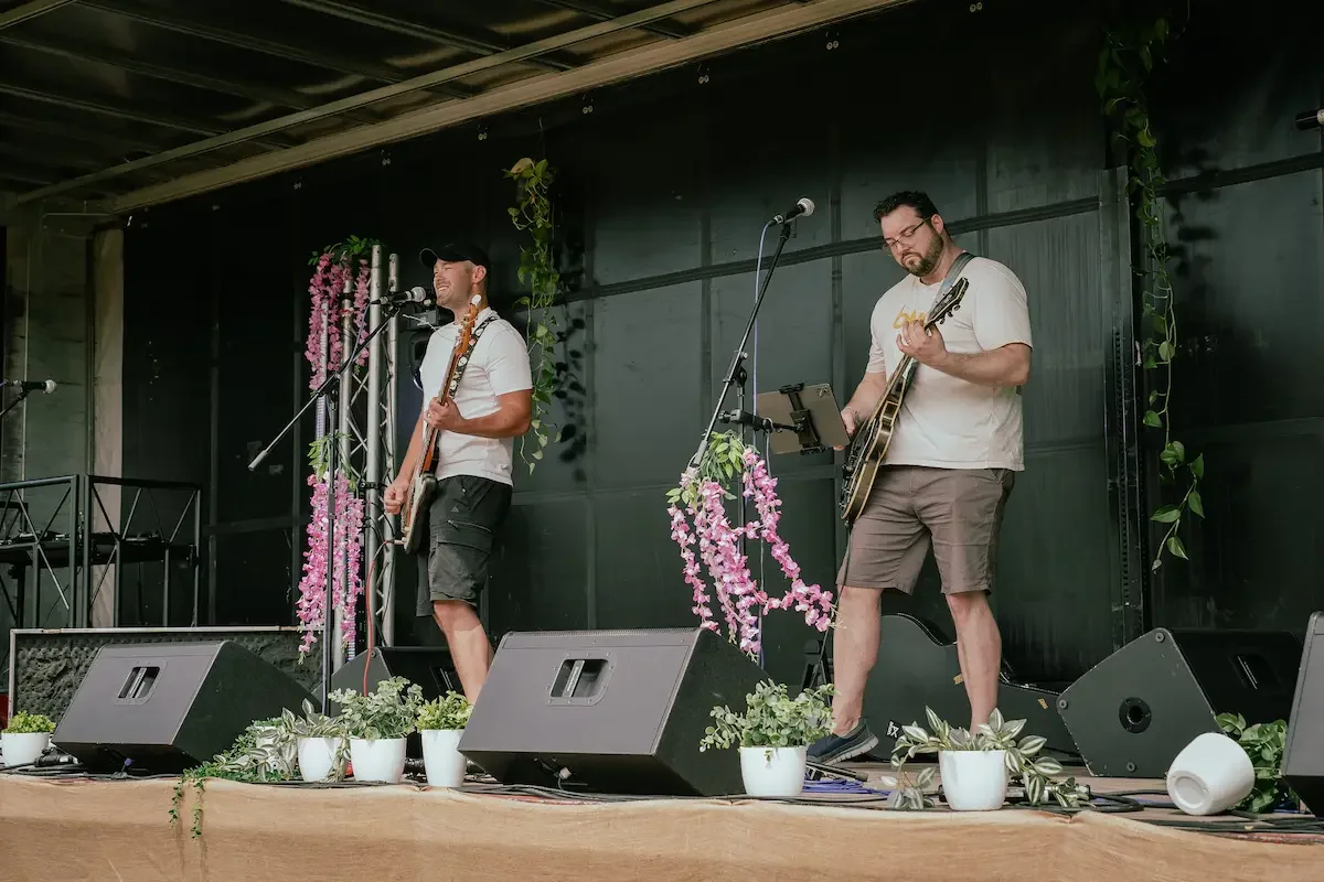 Two men on a stage with microphones and guitars performing at an outdoor summer gig