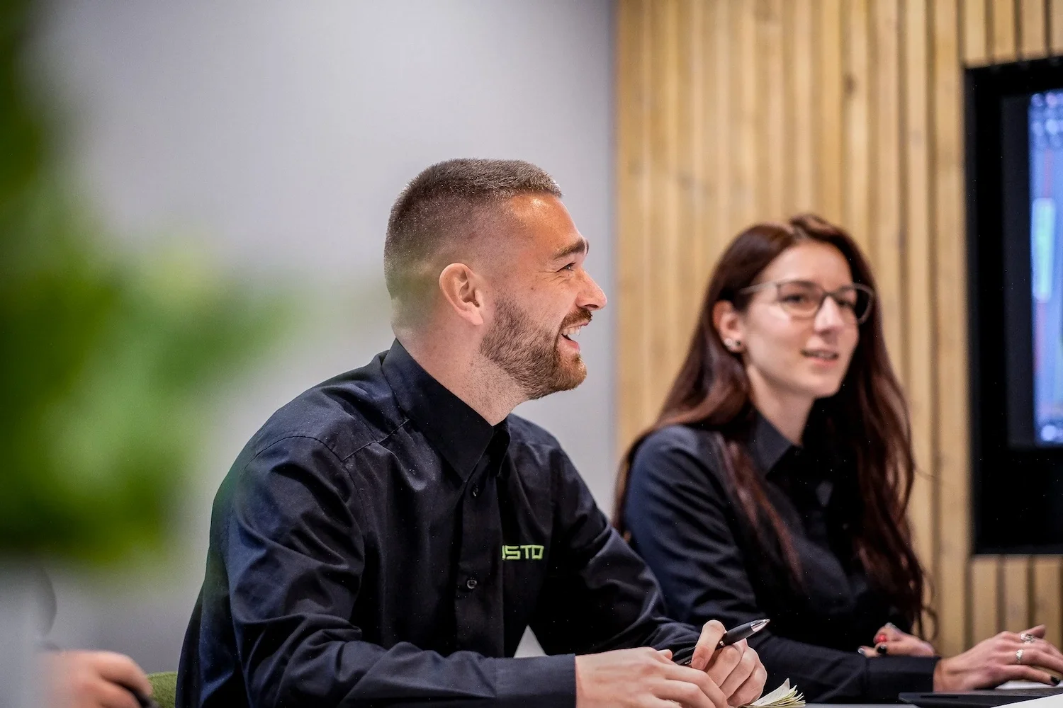 Two people, a man and a woman, sitting at a conference table, smiling and engaged in a discussion. The man has short hair and a beard, wearing a black shirt with green logo, holding a pen. The woman has long reddish-brown hair, glasses, and is also dressed in black, with a screen visible in the background.