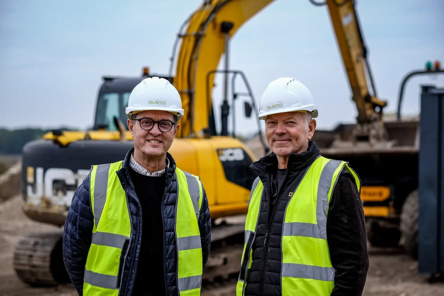 Two men, Steff & Jerome Wright on site stood in front of a digger. They are wearing hi vis and hard hats that say 'GUSTO'
