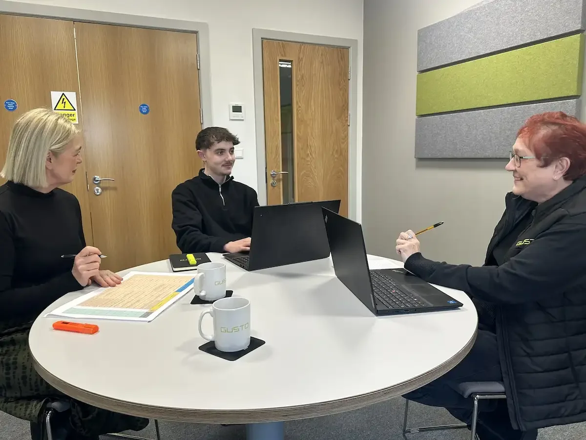 Two women and a man sat on a round table with notebooks and laptops having a meeting
