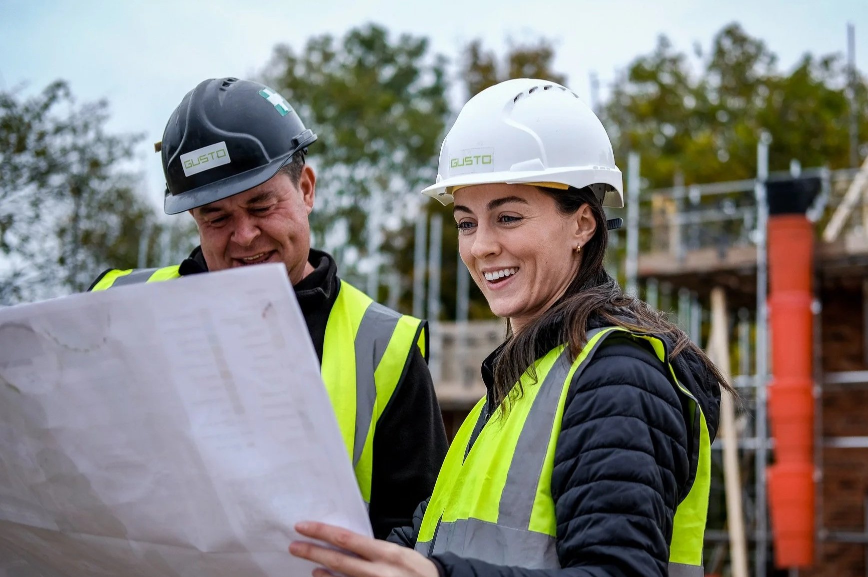 Construction site with a man wearing a hard hat and orange safety vest labeled 'GUSTO SITE MANAGER', looking at a blueprint or plan on a clipboard.