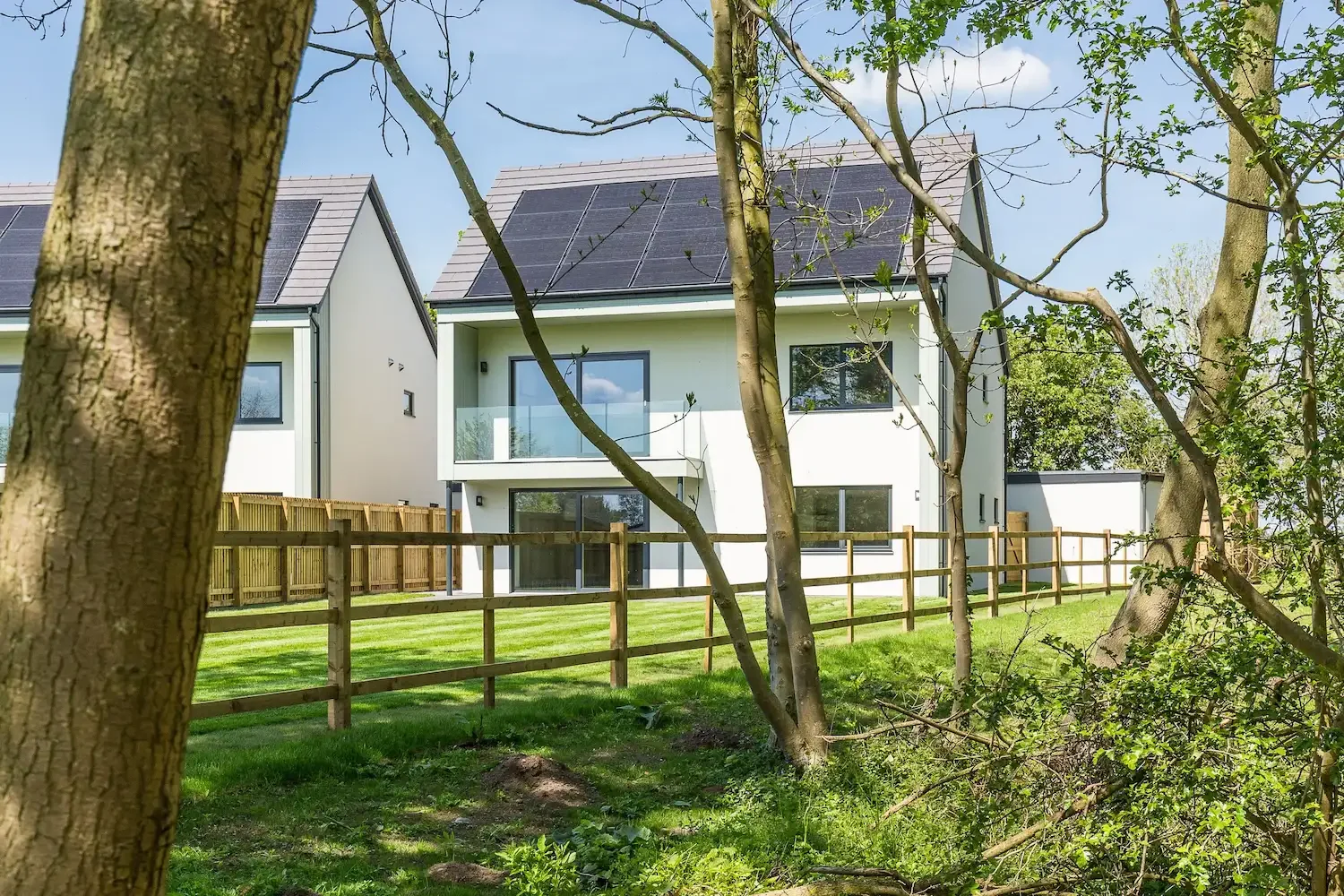 Modern white house with solar panels on the roof, large glass windows, and a balcony, surrounded by a wooden fence and green yard with trees. This house is at Woodlands Edge in Lincoln.