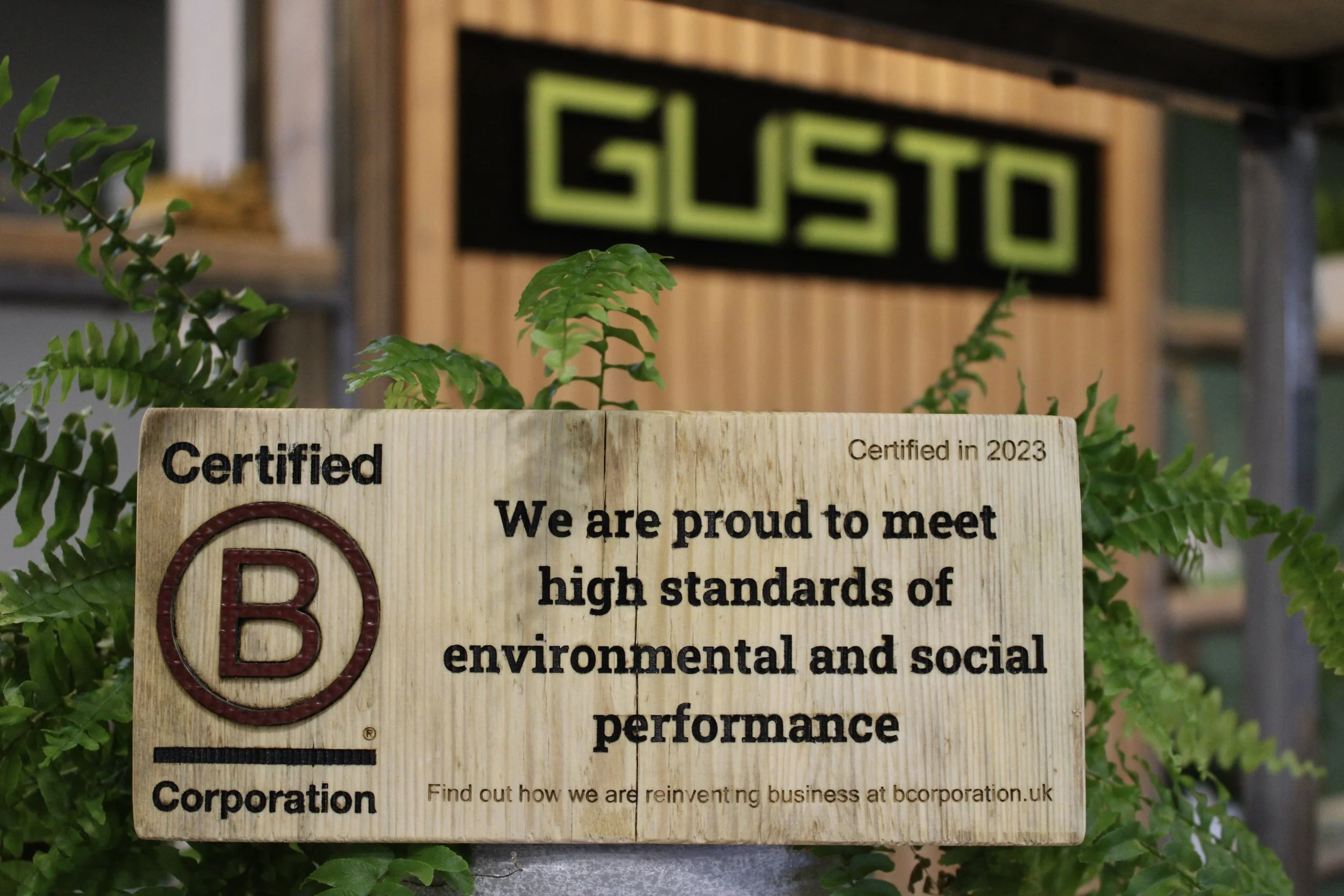 A close-up of a rustic wooden plaque engraved with the "Certified B Corporation" logo, noting Gusto Group met high standards of environmental and social performance in 2023.
