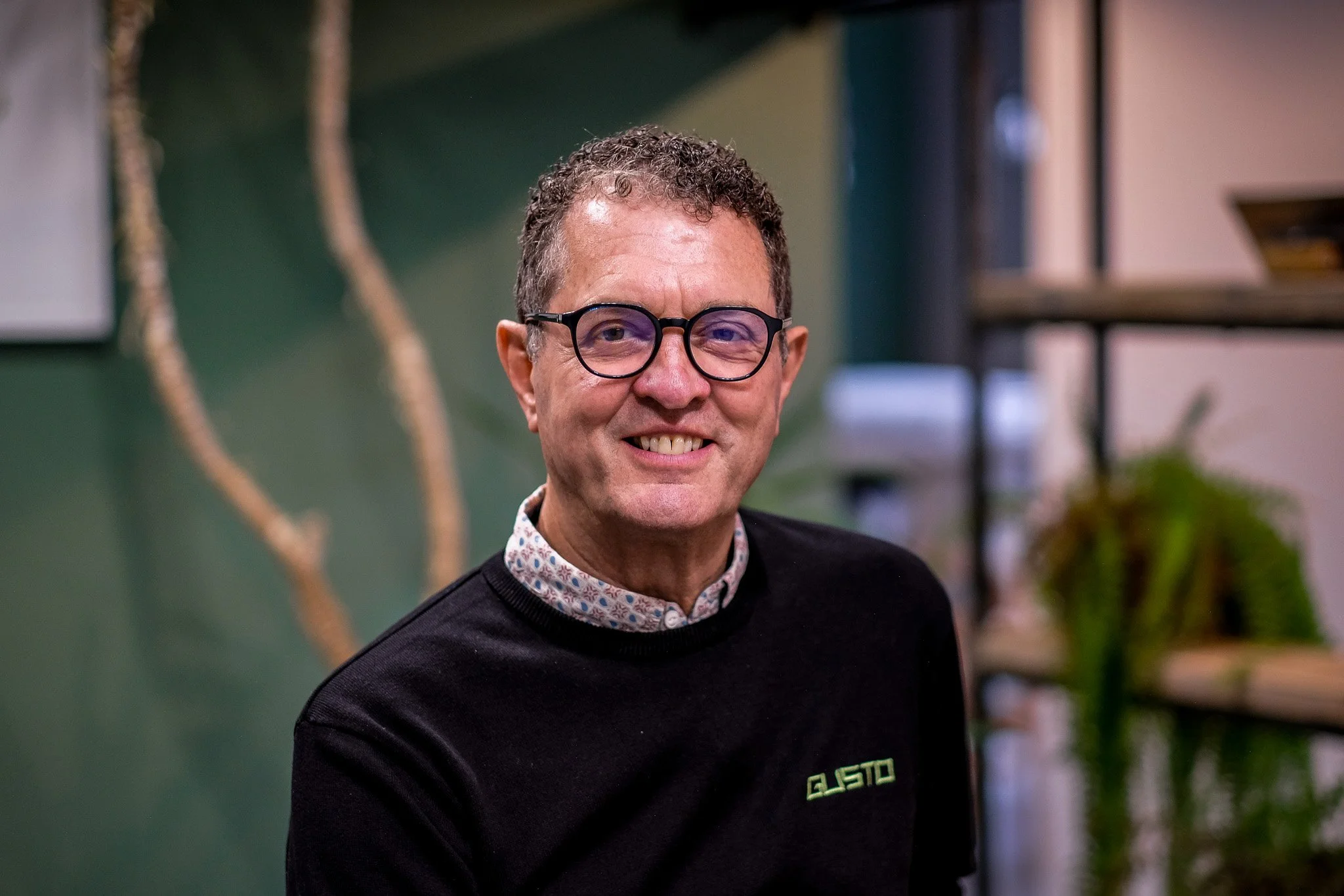 A middle-aged man with curly hair and glasses, wearing a checked shirt, standing indoors against a green and office background. Steff Wright