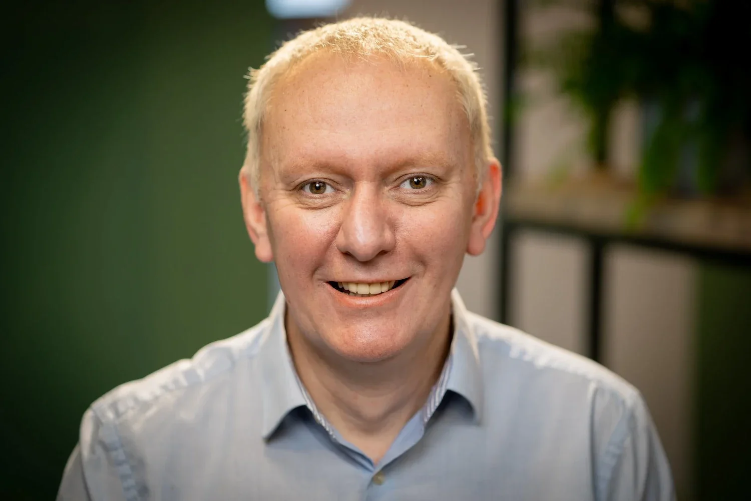 A smiling middle-aged man with short blond hair wearing a light gray collared shirt, indoor setting with blurred green background and shelves.
