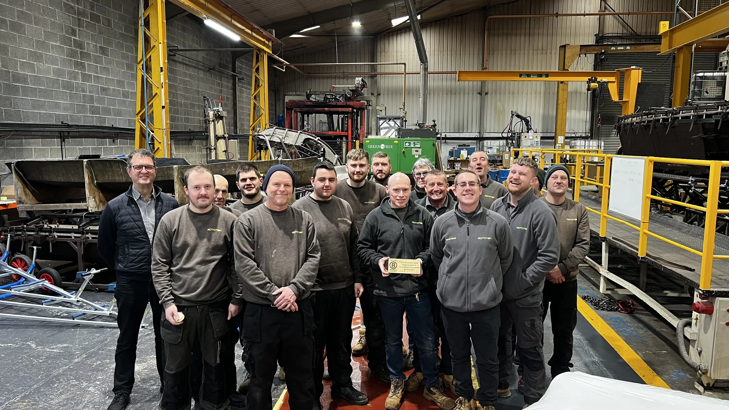 group of approximately 15 workers in grey Rototek uniforms standing inside a large manufacturing warehouse holding up a B Corp certification plaque
