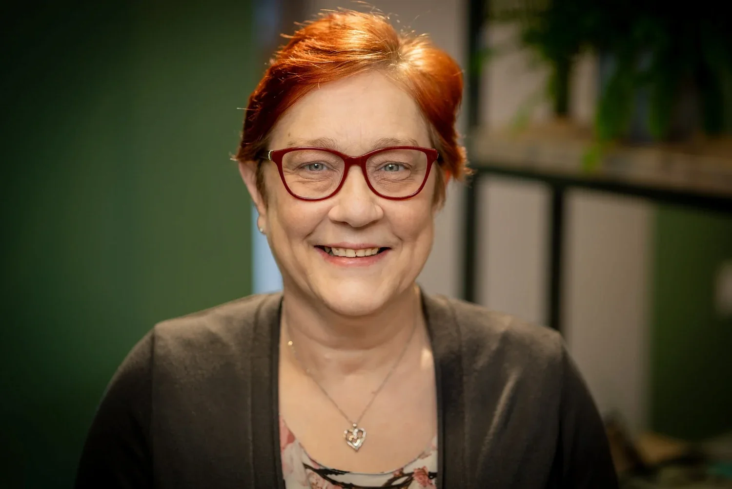 A smiling woman with short red hair, wearing glasses, a floral top, dark cardigan, and a heart-shaped necklace, sitting indoors with a blurred background of green and plants.