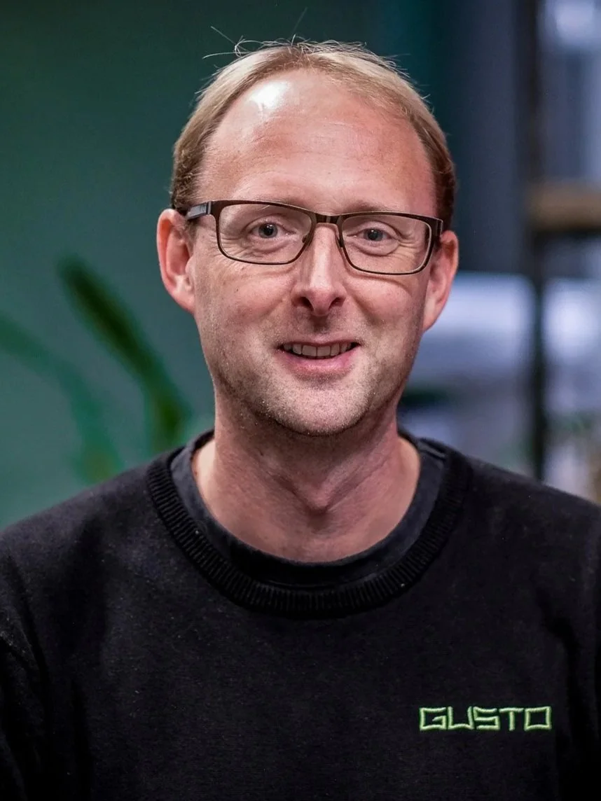 A man with glasses and light brown hair smiling, wearing a black shirt with the GUSTO logo embroidered on it, in an indoor setting.