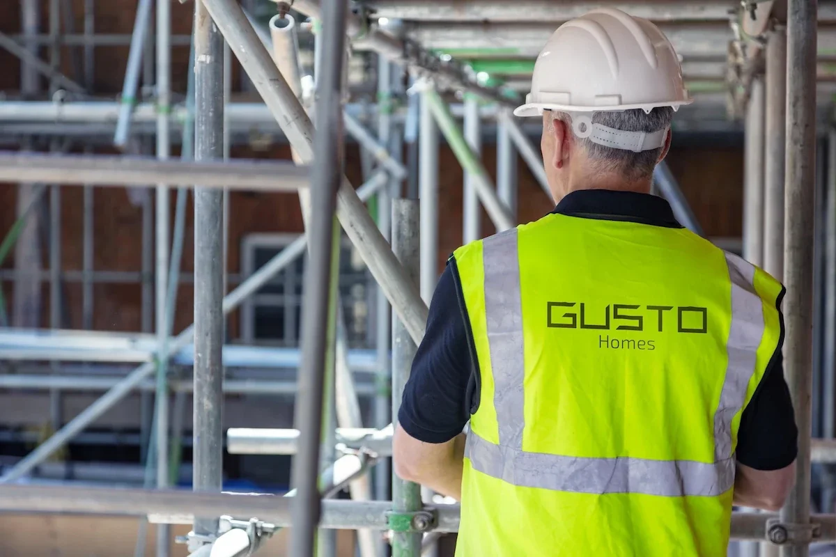 a man on a building site, walking across the scaffold, wearing a neon yellow hi vis, which says 'GUSTO Homes' and a white hard hat