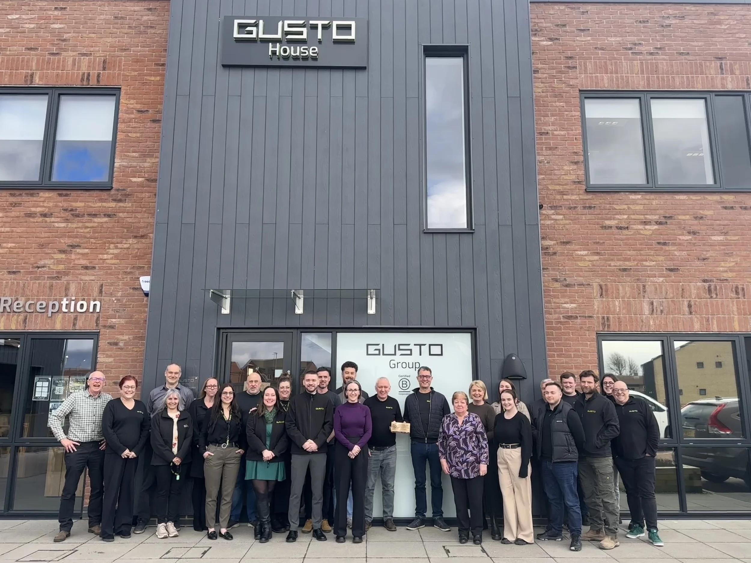 A large, diverse group of Gusto Group employees standing together and smiling in front of the modern brick and grey-paneled Gusto House building.
