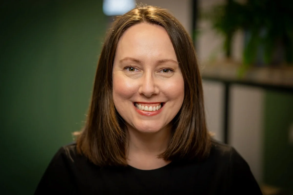 A woman with shoulder-length brown hair smiling at the camera, with a blurred green and indoor background.