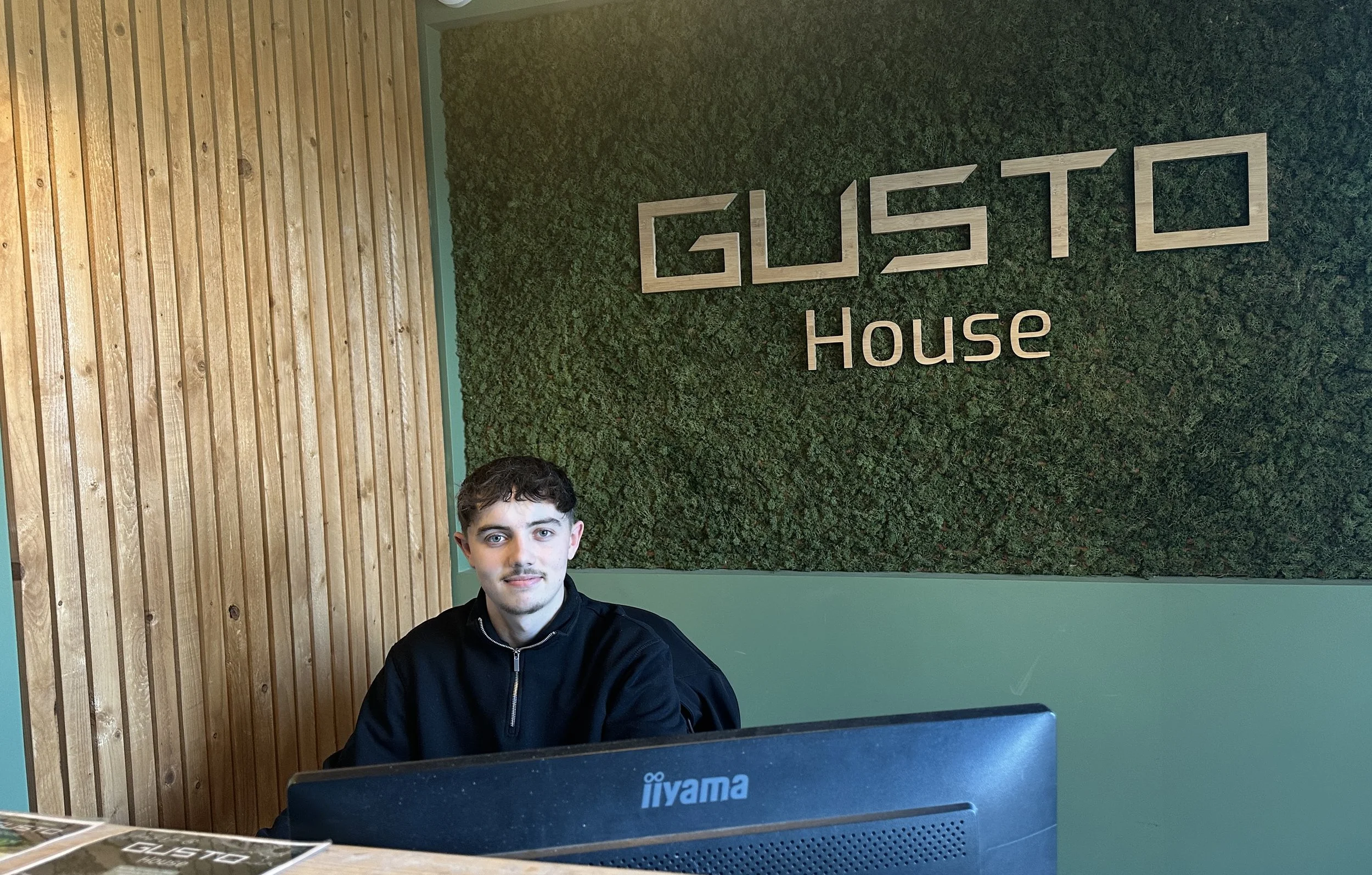 Tom smiling at the Gusto House reception desk in front of a branded moss wall.