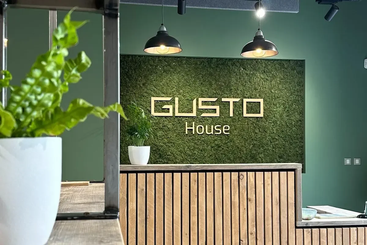 Indoor office space reception area with green wall, wooden slat counter, potted plant, green chair, and sign reading 'GUSTO House' with ceiling lights.
