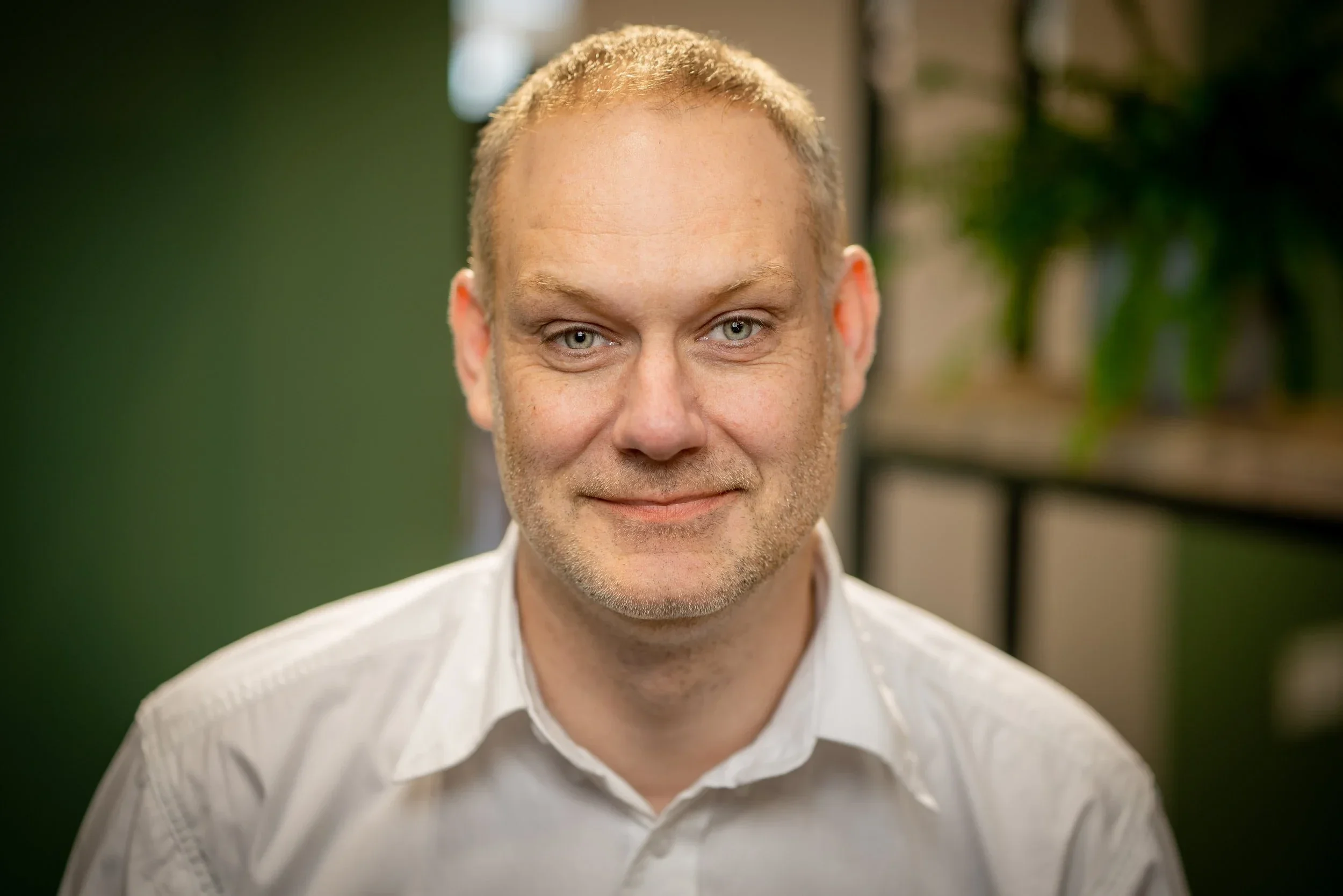 A man with short light-colored hair, blue eyes, and a slight smile, wearing a white shirt, sitting indoors with blurred background including green and dark elements.