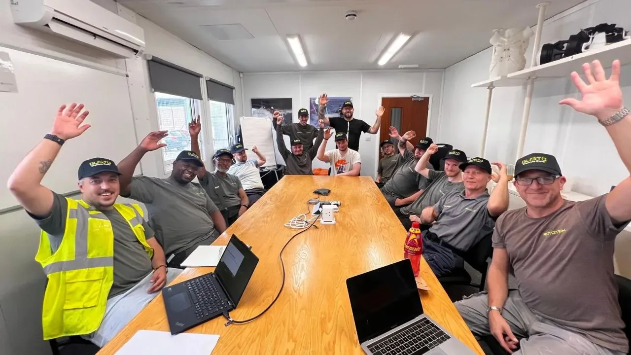 A group of men sat around a woodern table with their hands in the air wearing black baseball caps that say 'GUSTO Employee Owner'