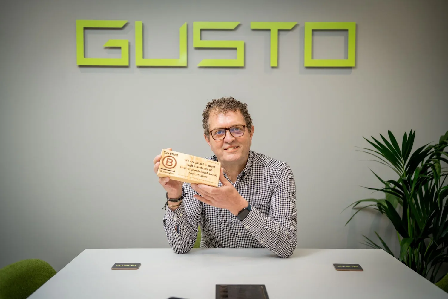 A middle aged man with curly hair and glasses sat at the head of a table in a boardroom, holding a wooden plaque that says 'certified B Corporation'