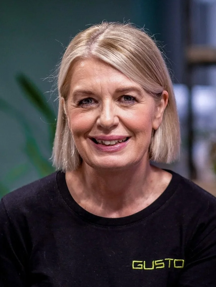 A smiling woman with blonde hair wearing a colorful, patterned blouse in an indoor setting with a green wall and plants