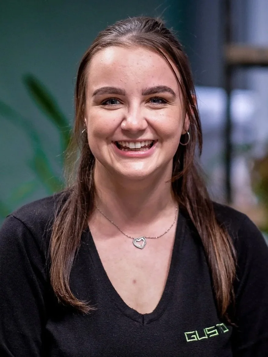 A young woman with dark brown hair, smiling, wearing a black and white striped shirt and a silver necklace with a heart-shaped pendant, standing in front of a green wall.