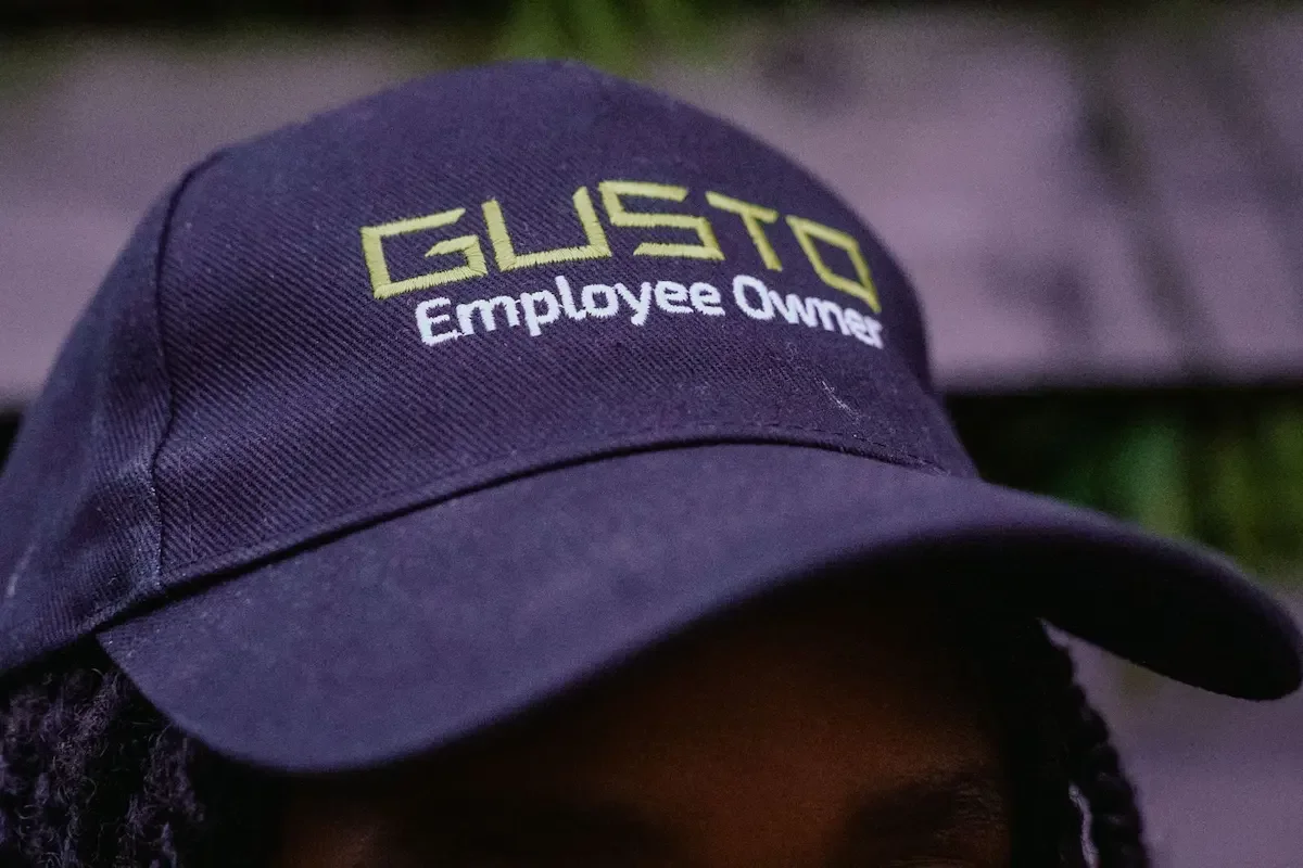 A close up of a person wearing a black baseball cap that says 'GUSTO Employee Owner'