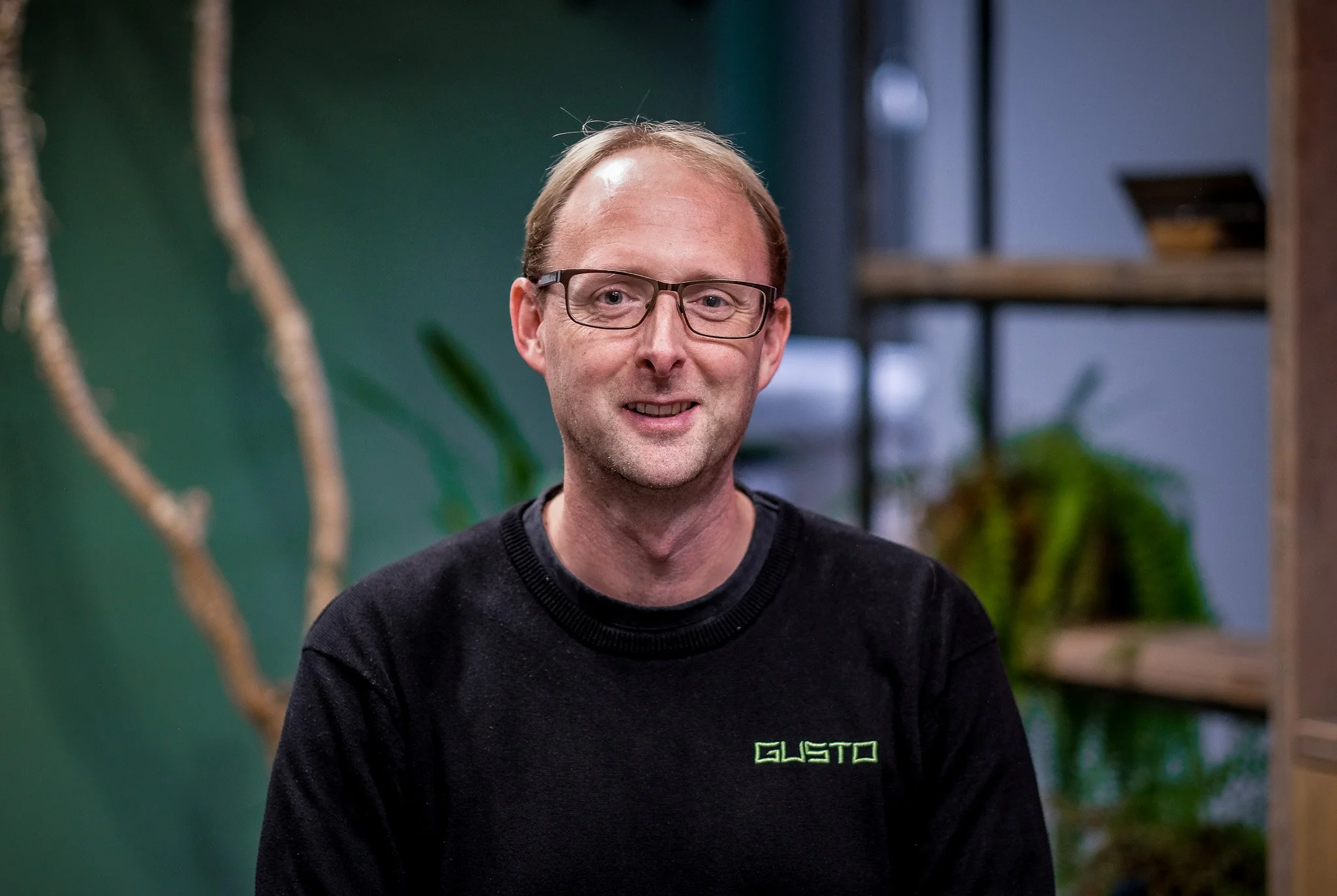 A man with glasses and light brown hair smiling, wearing a black shirt with the GUSTO logo embroidered on it, in an indoor setting.