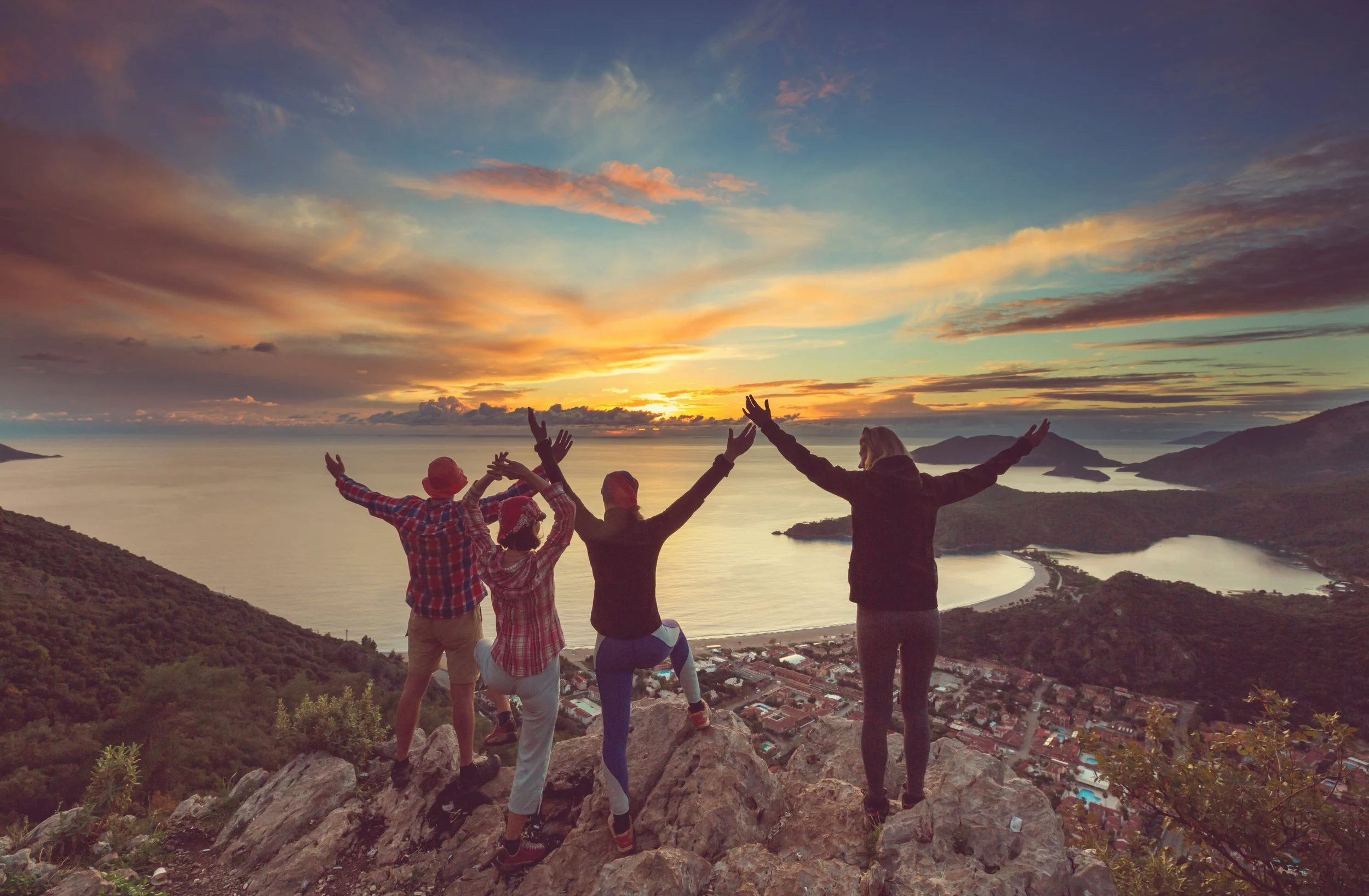 Four people standing on a rocky hilltop with arms raised, overlooking a body of water and a small town at sunset.