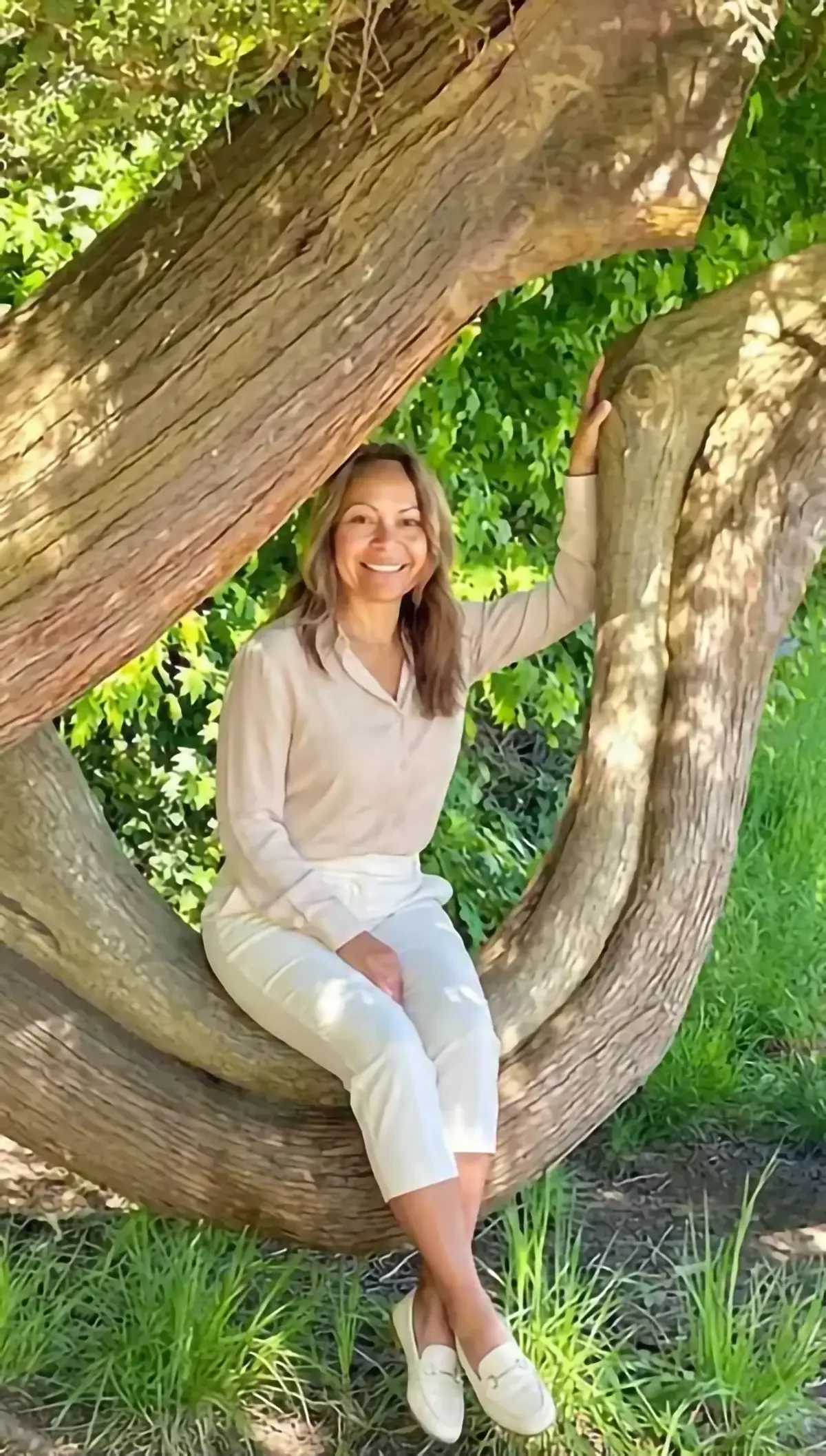 A woman sitting on a large tree branch, smiling, surrounded by green leaves and grass in a sunny outdoor setting.