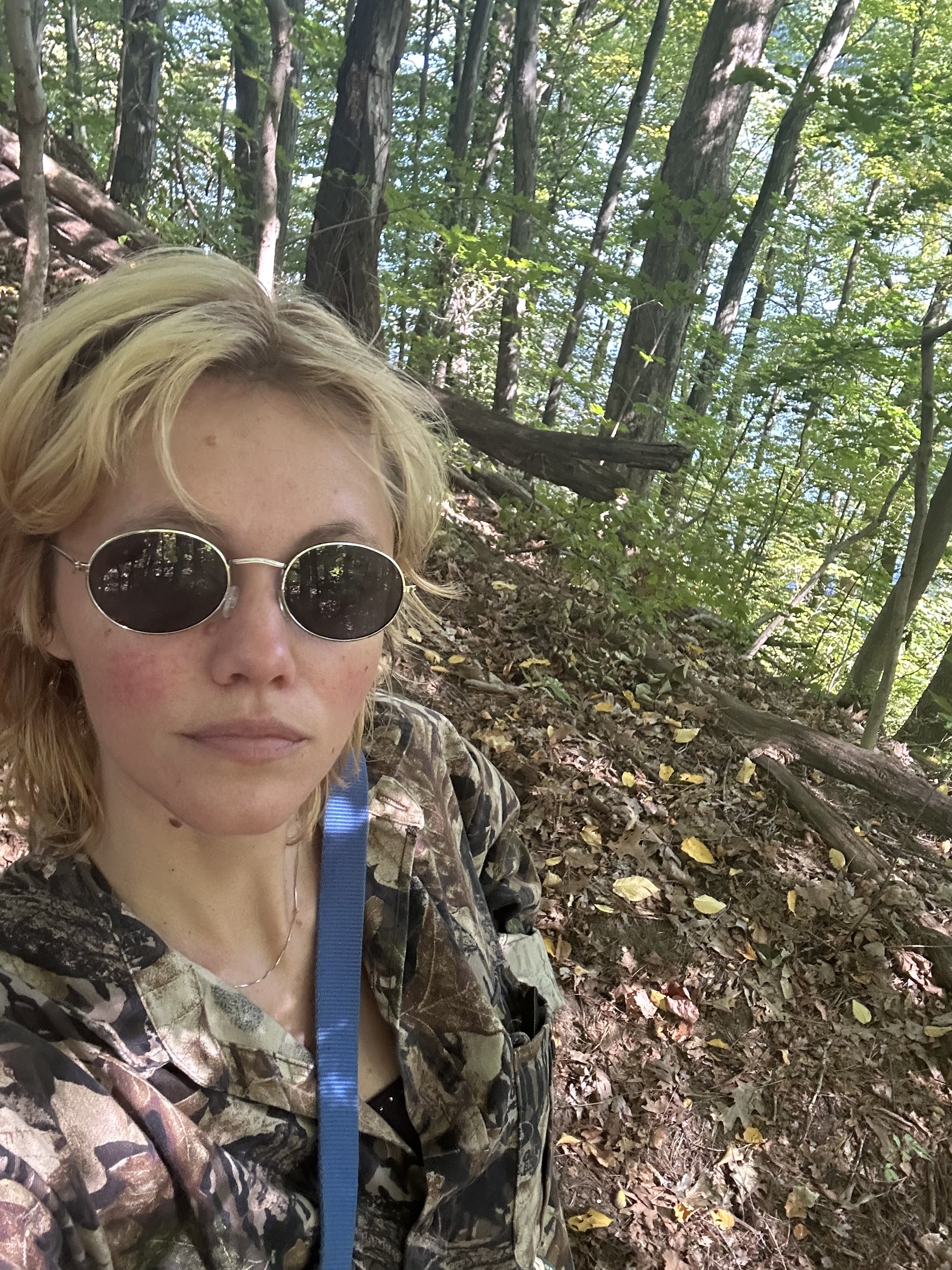 Blonde white person with short hair and sunglasses self in the woods.