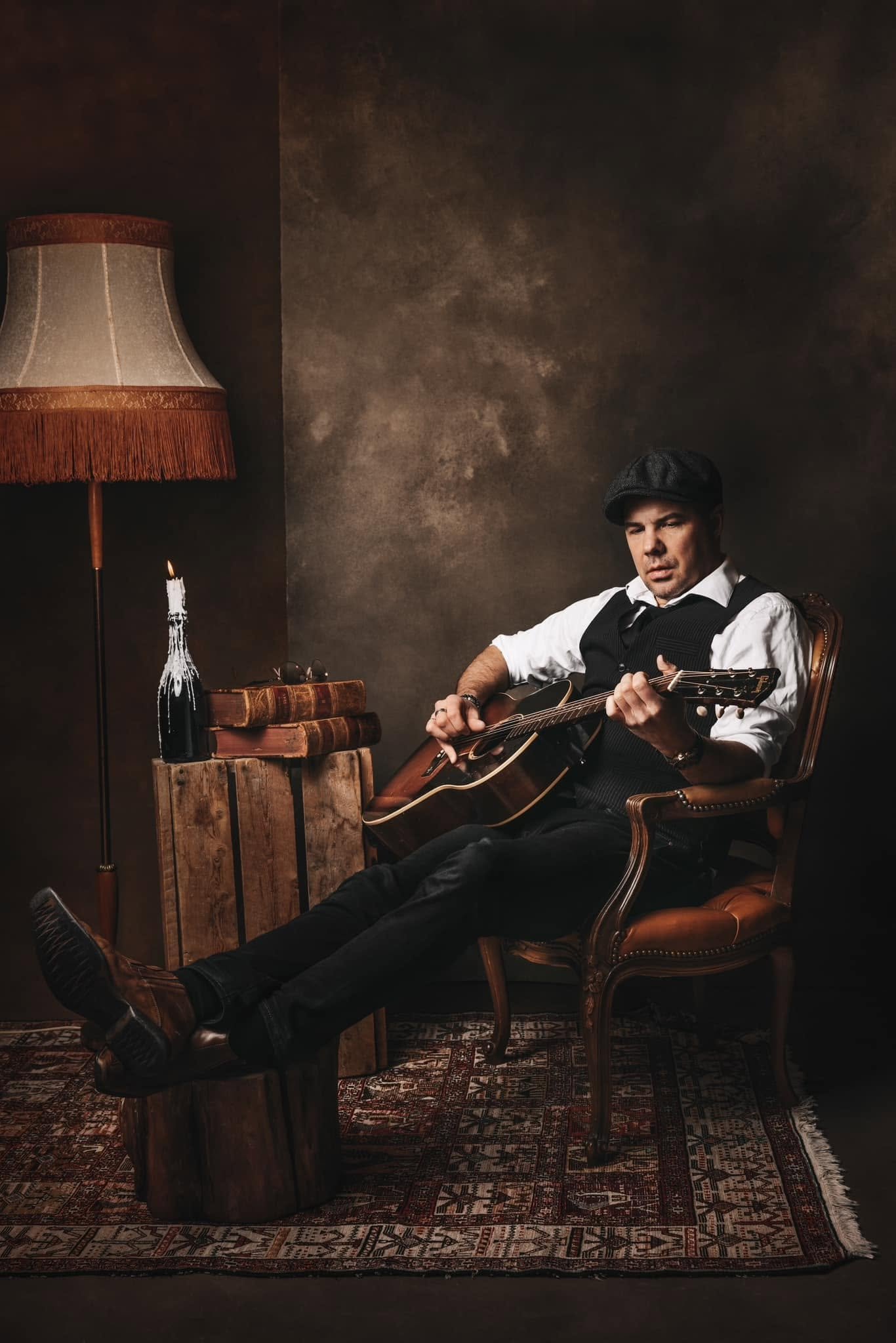 En mann som spiller akustisk gitar, sitter i en komfortabel lenestol. Han har på seg en flat caps, hvit skjorte, svart vest og svarte bukser. Bak ham er det en vintage-stil klokkestokk, et bord laget av trær med bøker, en brennende stearinlys, og en eldre lampe med en brun skygge. Rødbrunt teppe dekker gulvet.