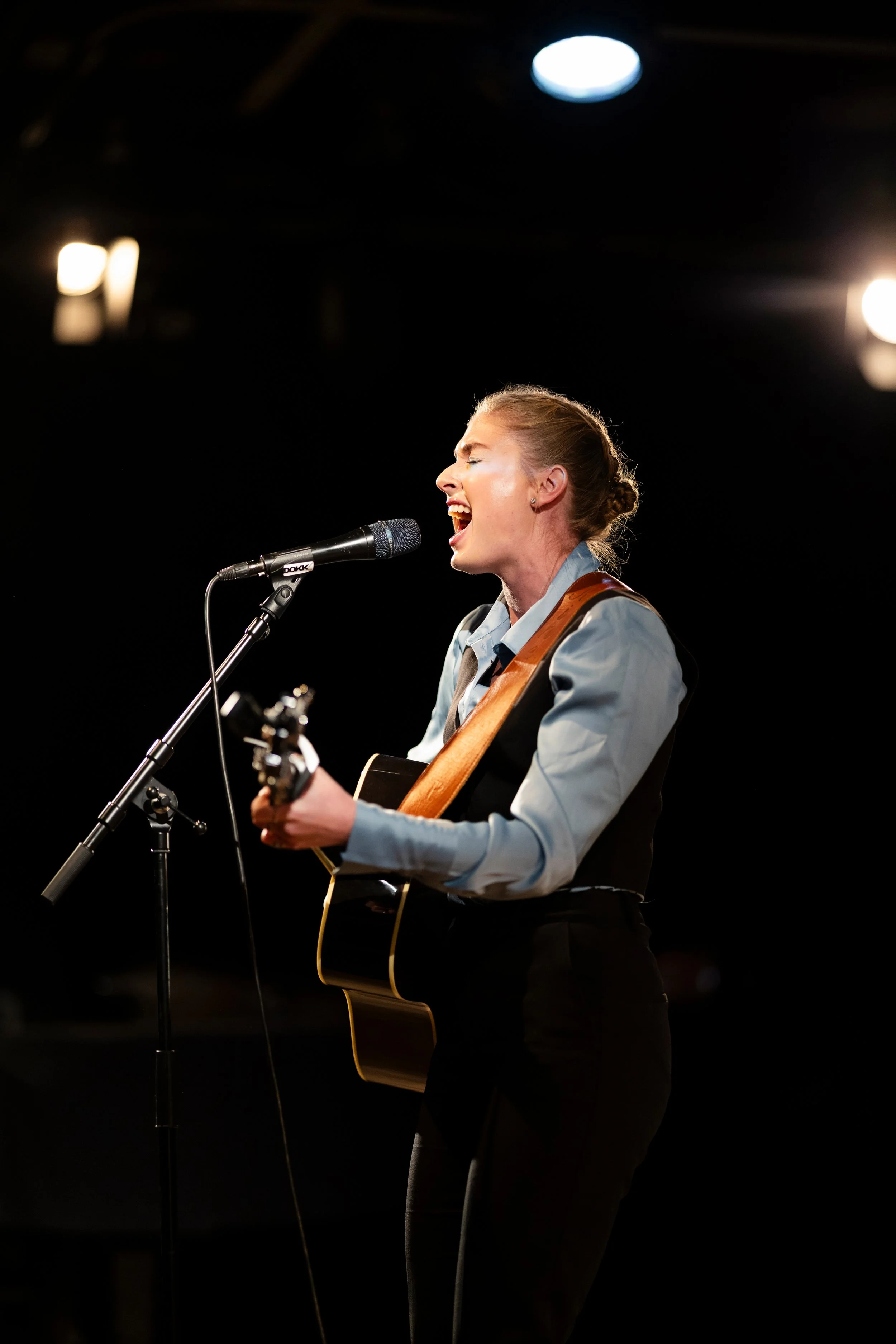 En kvinne som synger med mikrofon og spiller gitar under en konsert, bakgangen er mørk med lyspærer og tak. 