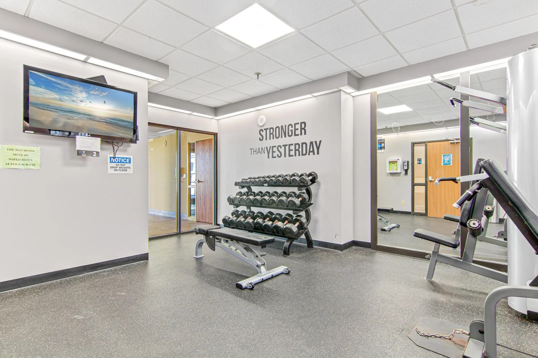 A fitness center with a television mounted on the wall, a rack of dumbbells, a workout bench, and various exercise equipment in a bright room with white ceiling tiles and motivational wall art reading 'Stronger Than Yesterday'.