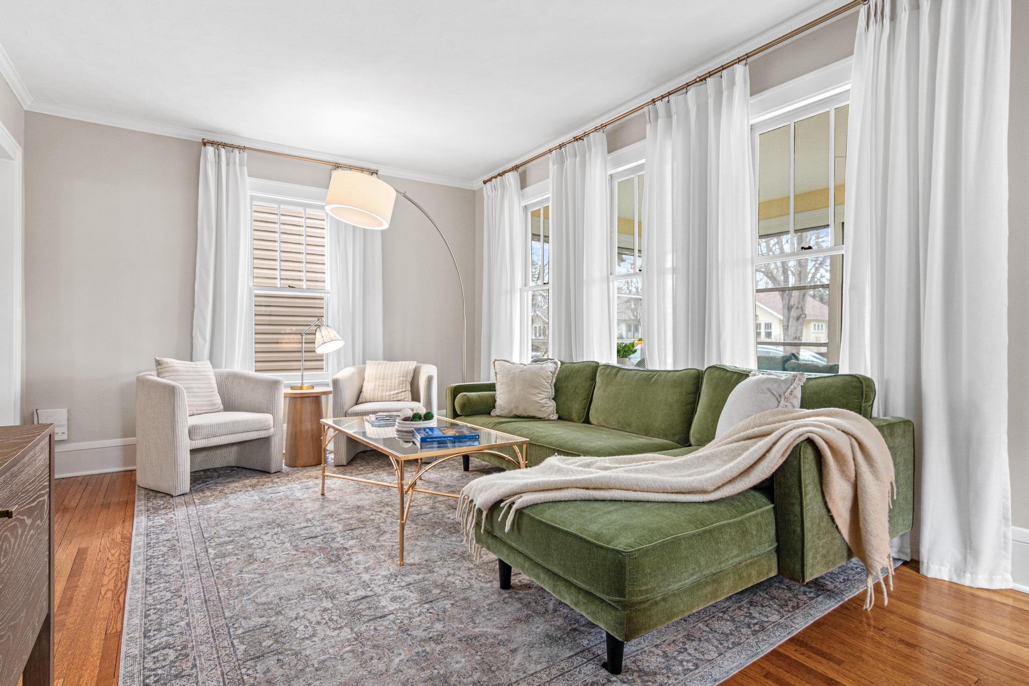 Bright living room with three large windows, a green sectional sofa, two cream armchairs, a wooden side table, a glass top coffee table, a floor lamp with a large white shade, white curtains, and a patterned area rug.