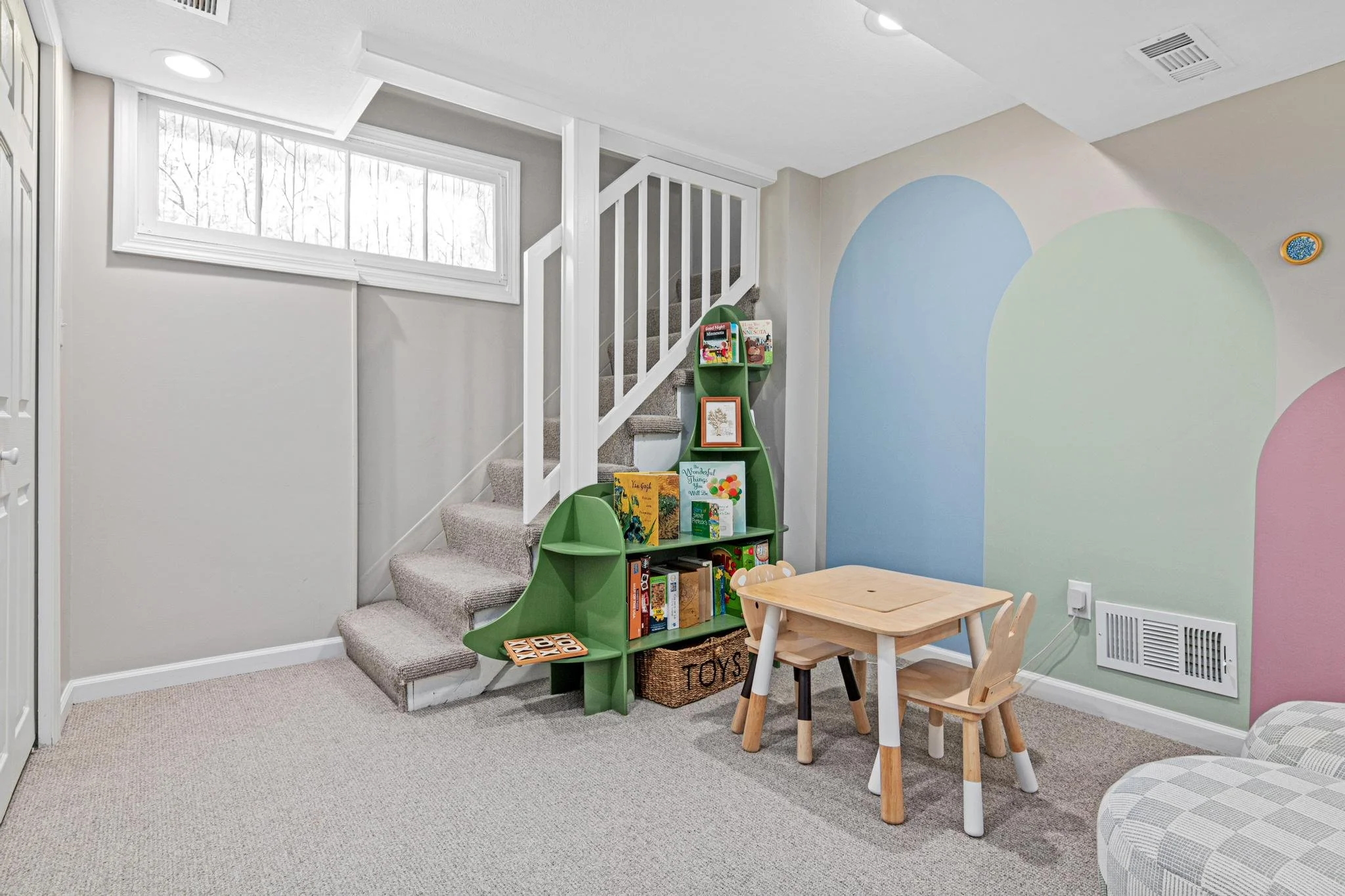 Interior of a children's playroom or corner with a small wooden table and chairs, a colorful bookshelf shaped like a tree, and wall painted with pastel arches. There's a window near the staircase, and a plush armchair is partially visible.