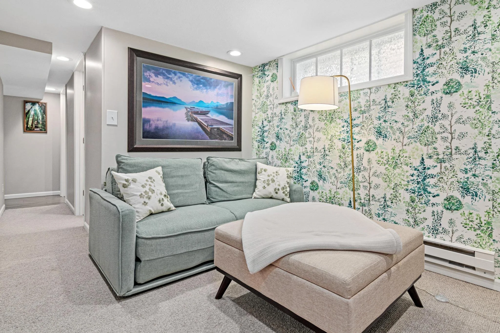 A cozy living room corner with a light green sofa, decorative pillows, a beige ottoman with a throw blanket, a tall floor lamp, floral wallpaper, a large framed landscape painting, and a window with white trim.