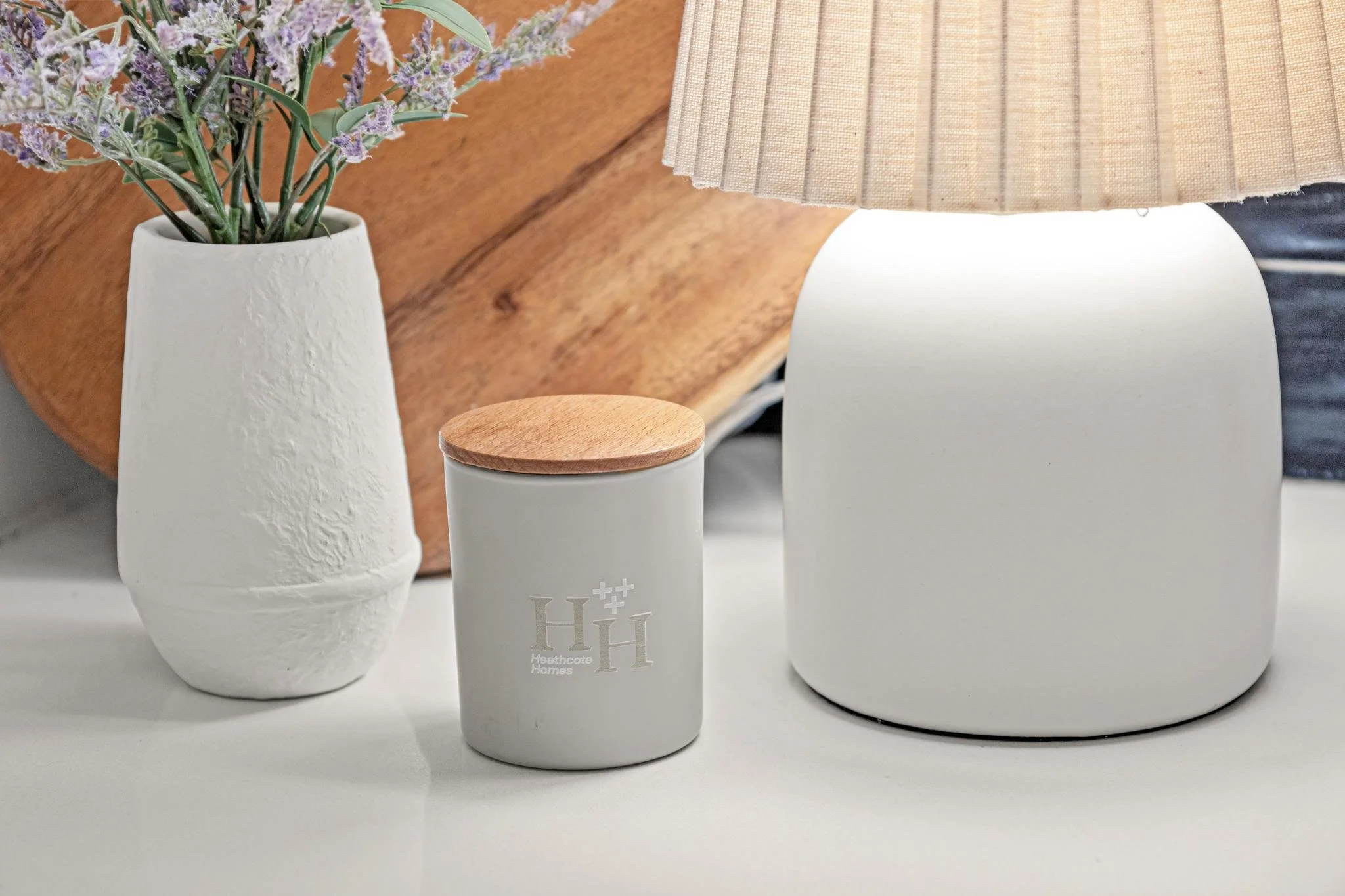 A white vase with a textured surface holding purple flowers, a white container with a wooden lid and medical symbols, and a white table lamp with a beige striped lampshade on a white surface.