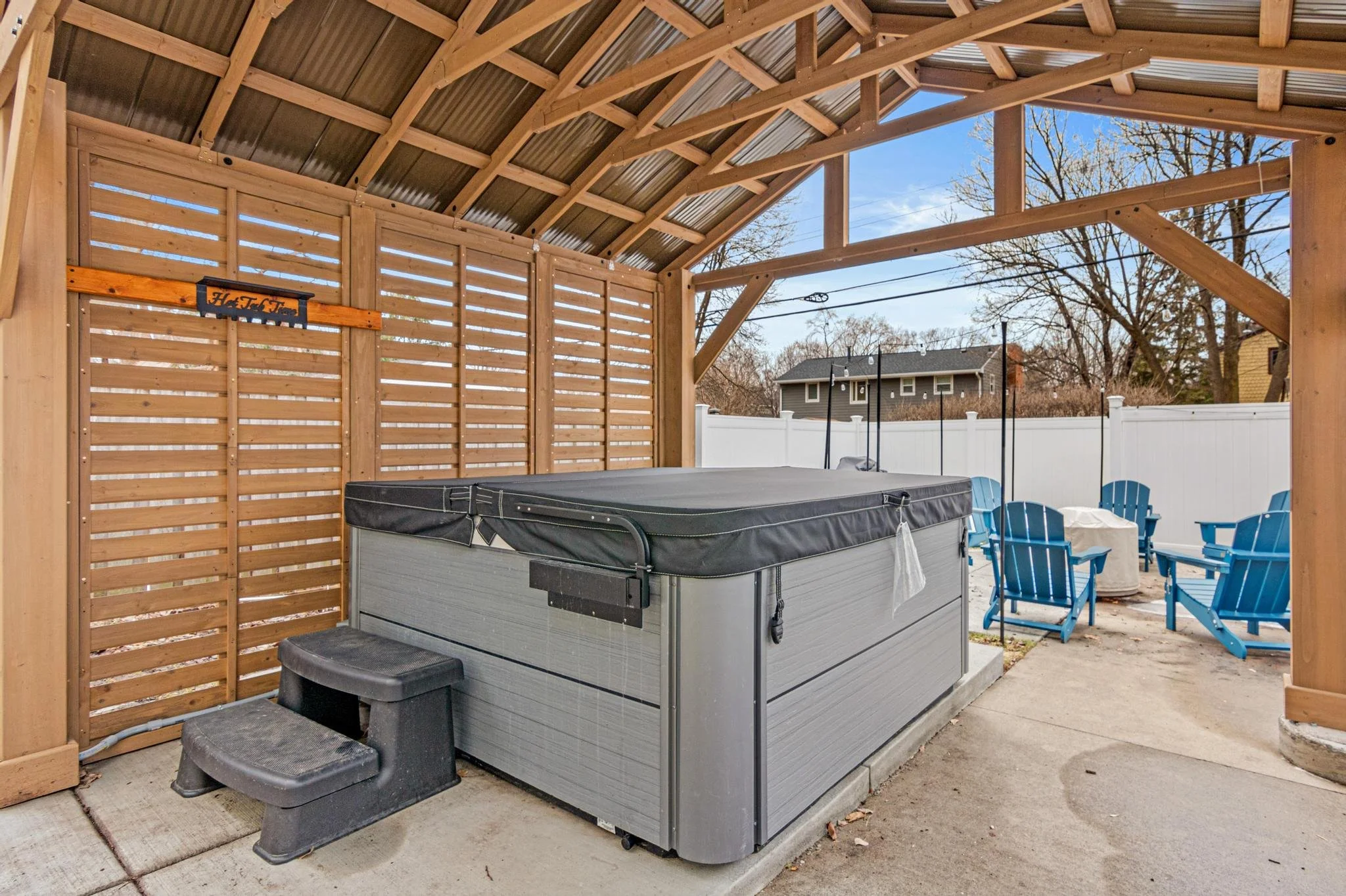 An outdoor hot tub in a wooden shelter with a slatted wall, a small black step stool, and a seating area with four blue Adirondack chairs and a fire pit.