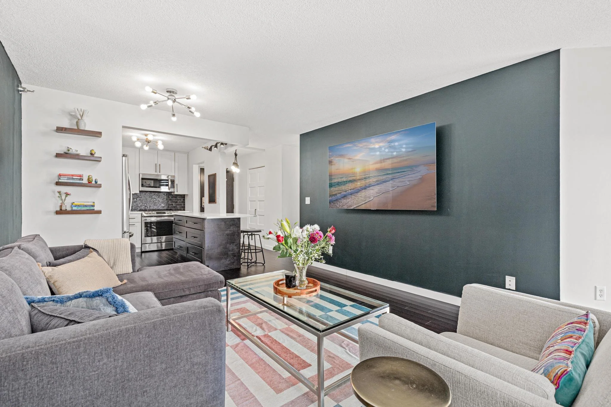 Living room with gray sectional sofa, a glass coffee table with a flower arrangement, and a large TV on a dark accent wall showing a beach sunset, with a nearby open kitchen with a dark island and white cabinets.