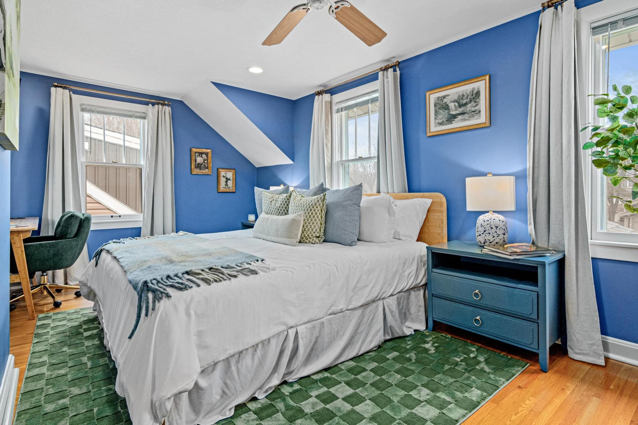 A bedroom with blue walls, a large bed with white linens and multiple decorative pillows, a green patterned area rug, a blue nightstand with a lamp and magazines, a window with white curtains, and framed artwork on the walls