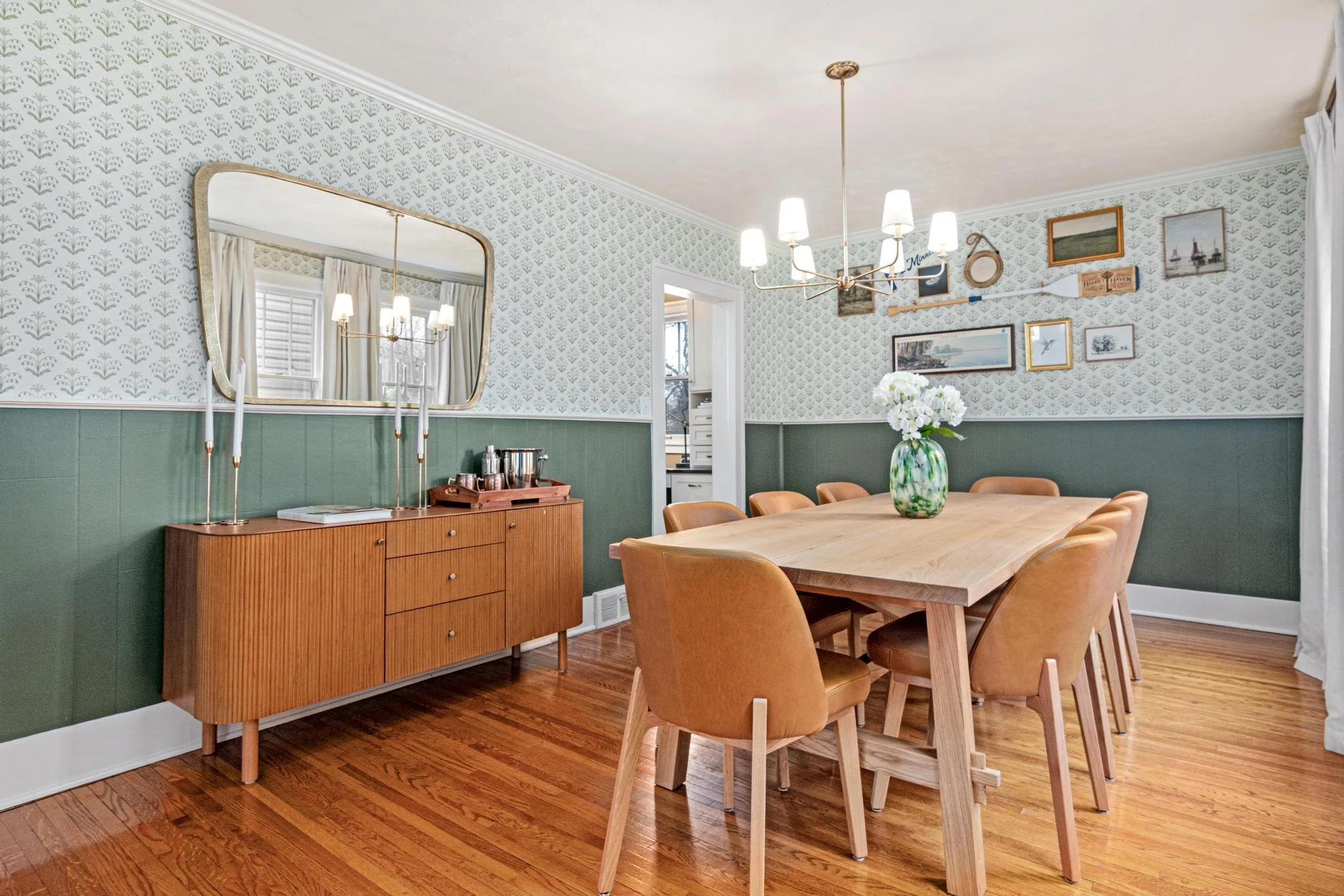 A dining room with a wooden table and tan chairs, a wooden sideboard with a tray of metalware, a large mirror, a chandelier, and a vase of white flowers, featuring green and white wallpaper and hardwood floors.