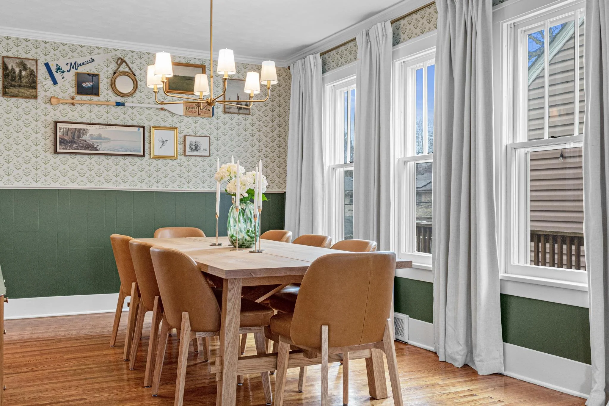 A dining room with a wooden table and six tan chairs. There is a green and white floral vase with white flowers and candles on the table. The room has large windows with white curtains, beige wallpaper with a green wainscot, and a chandelier hanging 