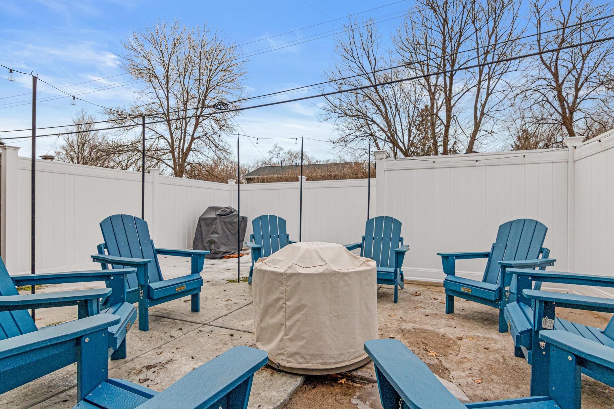 Backyard patio with six blue Adirondack chairs arranged in a circle around a covered table, a grill covered with a black cover, white fencing, leafless trees, and a clear blue sky.