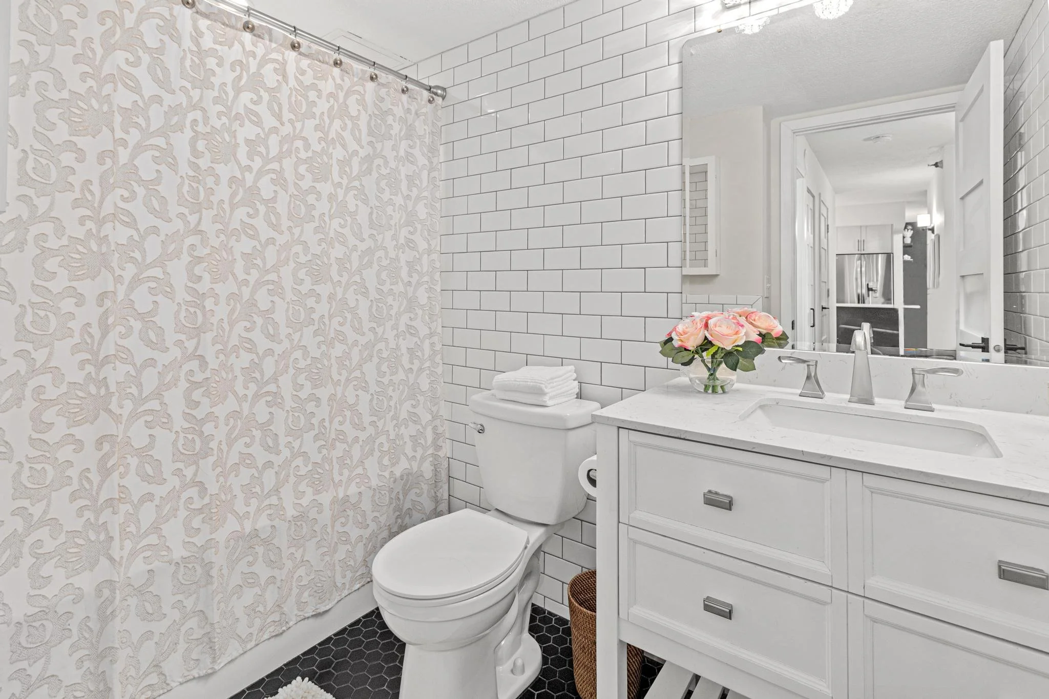A modern bathroom with white subway tile walls, a white toilet with a stack of white towels on top, a vanity with a white countertop, a vase of pink roses, a large mirror, a decorative shower curtain, and black hexagonal floor tiles.