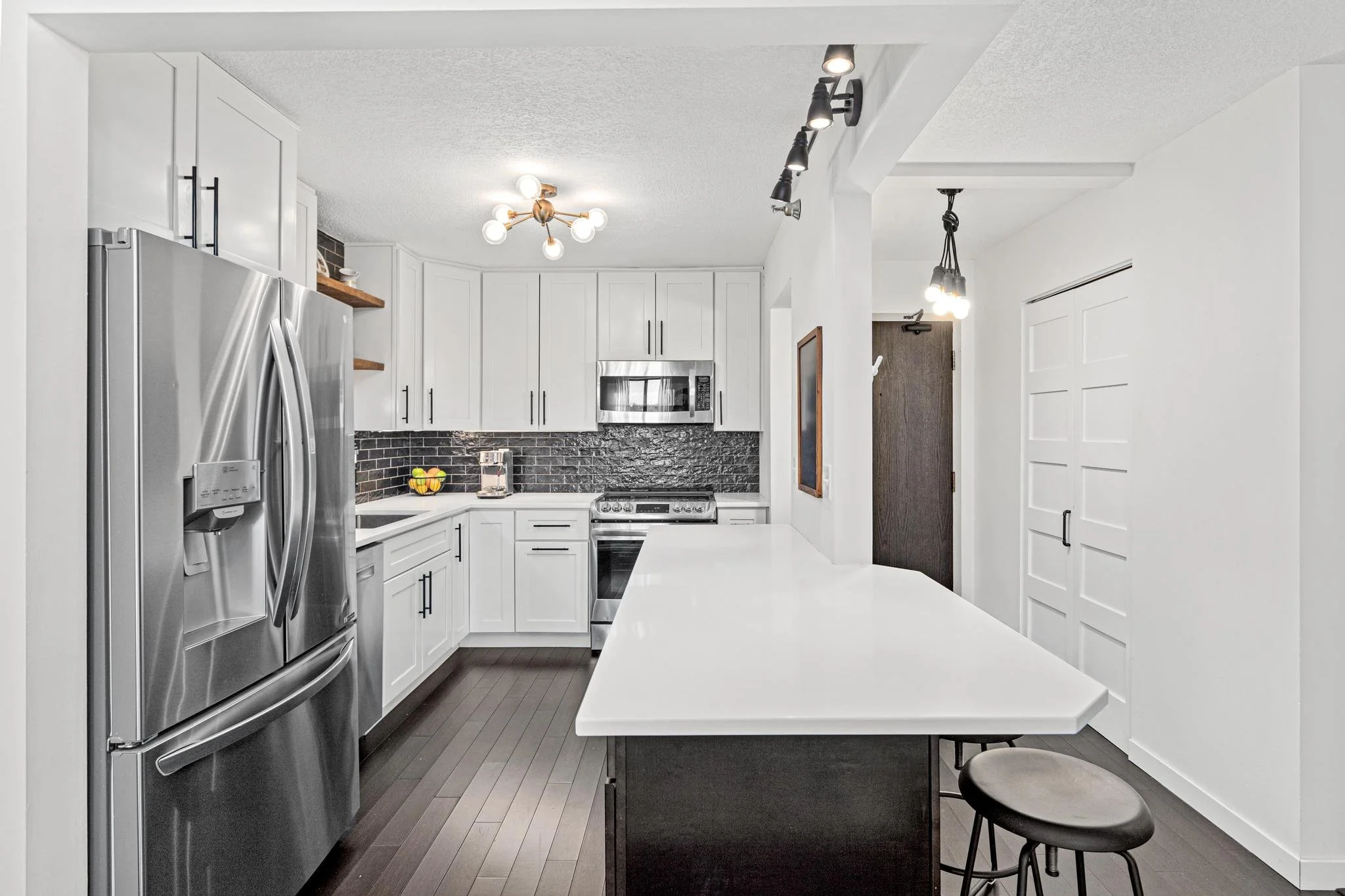 Modern kitchen with white cabinets, black backsplash, stainless steel appliances, dark hardwood floors, and a white countertop island with bar stools.