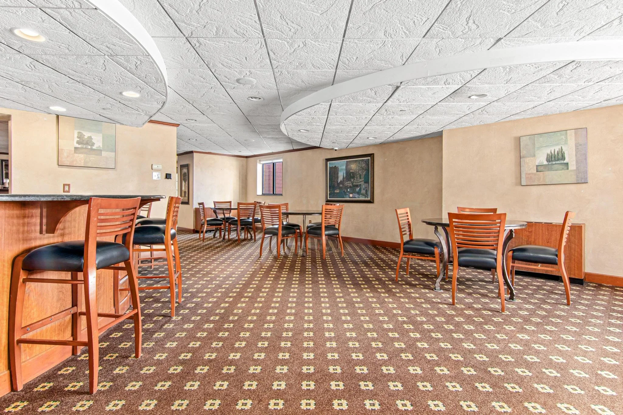 Empty dining area with wooden chairs with black cushions and patterned carpet. Beige walls with paintings, small windows, and a curved ceiling.