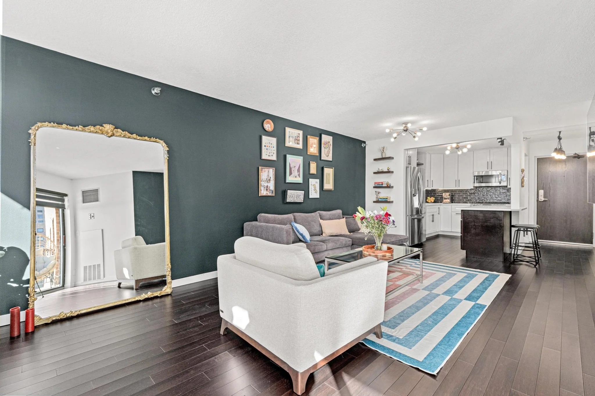 Living room with dark hardwood floors, a large ornate mirror reflecting a window and chair, a dark green accent wall with picture frames, a gray sectional sofa, a white armchair, a multicolored striped area rug, a coffee table with a vase of flowers,