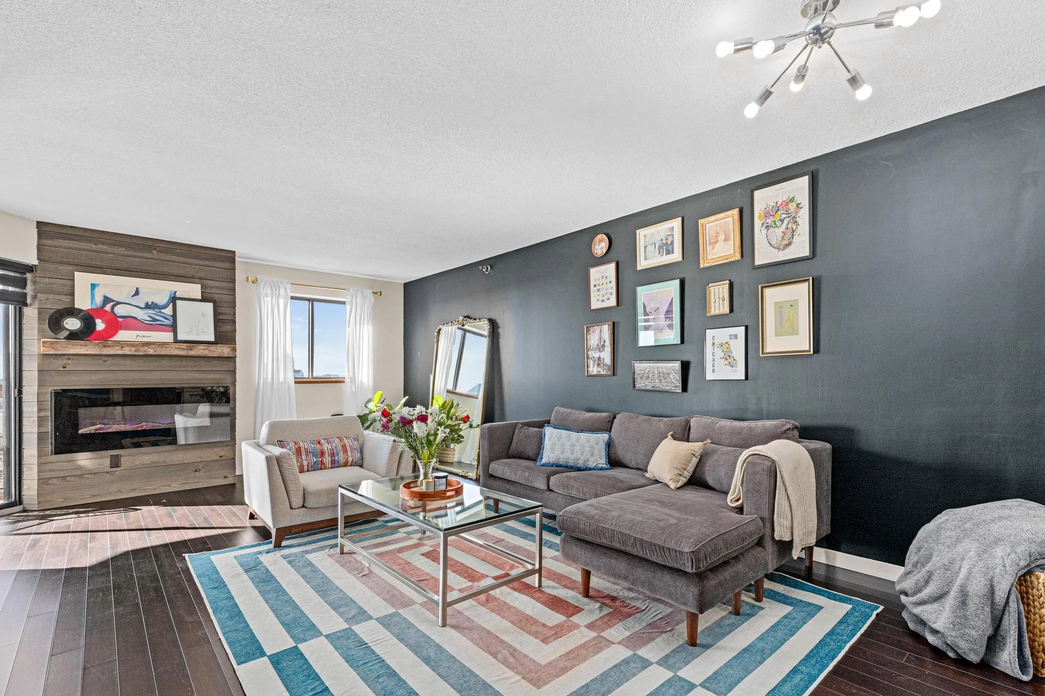 Living room with grey sectional sofa, white armchair, colorful geometric area rug, black accent wall with gallery wall of framed art, mirror, and clock, fireplace with wooden surround, window with white curtains, modern ceiling light fixture, and a c