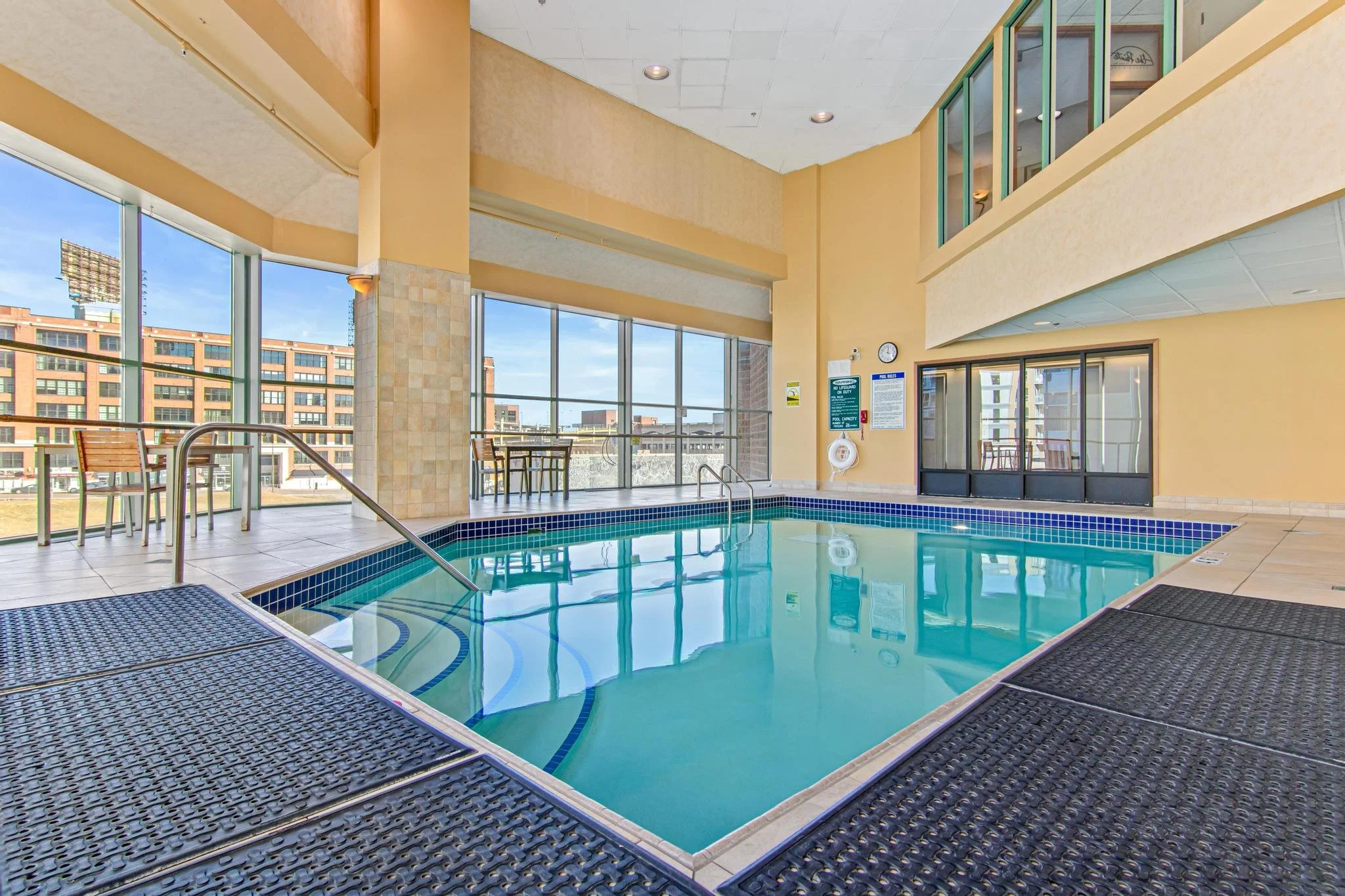 Indoor swimming pool with large windows, metal handrails, tiled edges, and a seating area with chairs and tables, in a modern building.
