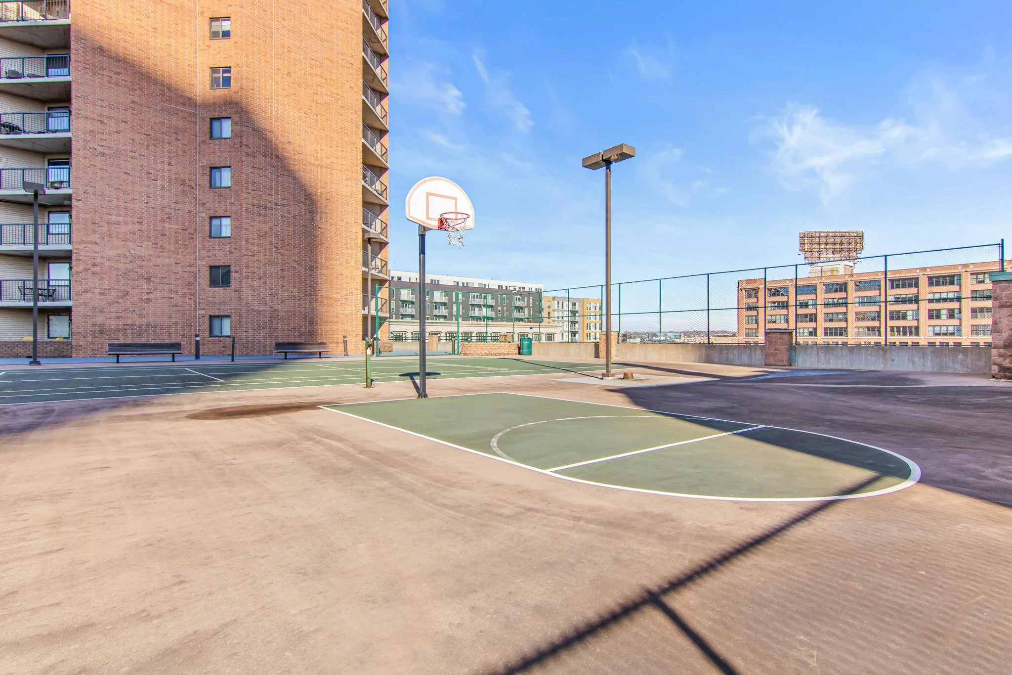 An outdoor basketball court on a rooftop with a brick apartment building on the left, surrounded by tall buildings and a clear blue sky.