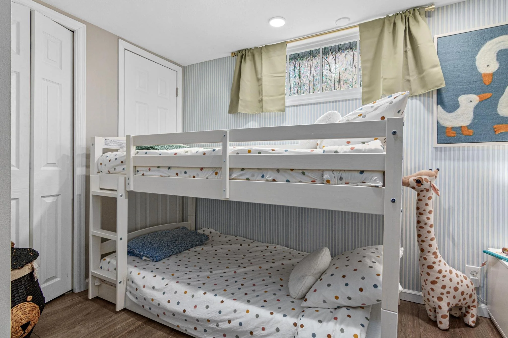 A children's bedroom with a white bunk bed and polka dot bedding, a plush giraffe toy, a window with beige curtains, and wall art of ducks.