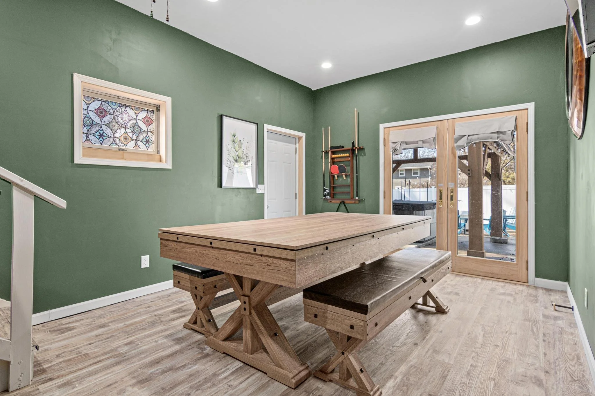 A dining room with green walls, a wooden table with benches, sliding glass doors, decor, and a pool visible outside.